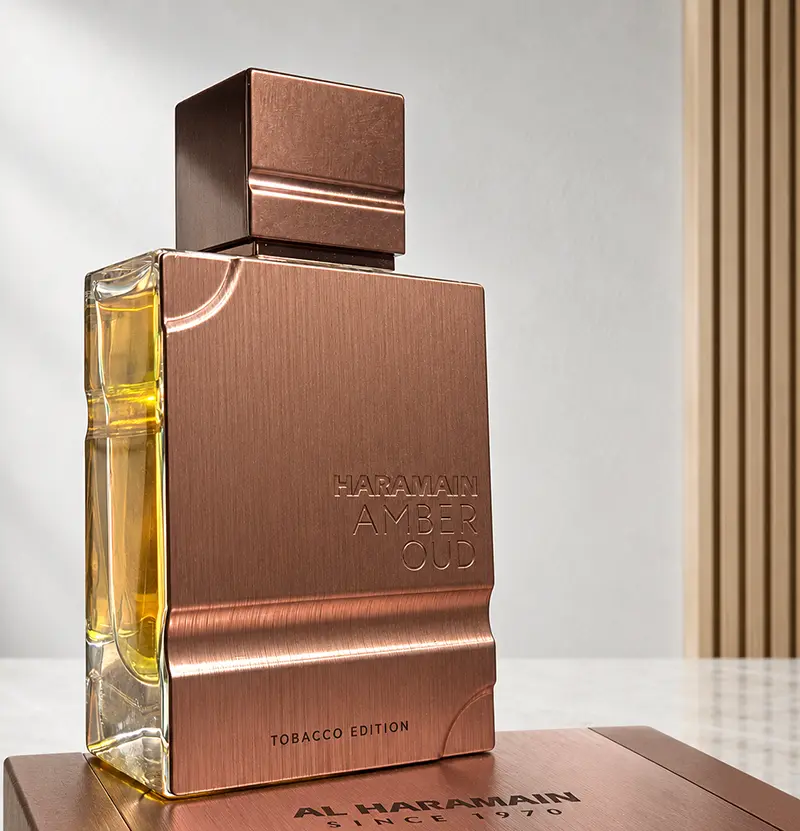 Angled Al Haramain Amber Oud Tobacco Edition bottle on its box, showing the clear side glass.