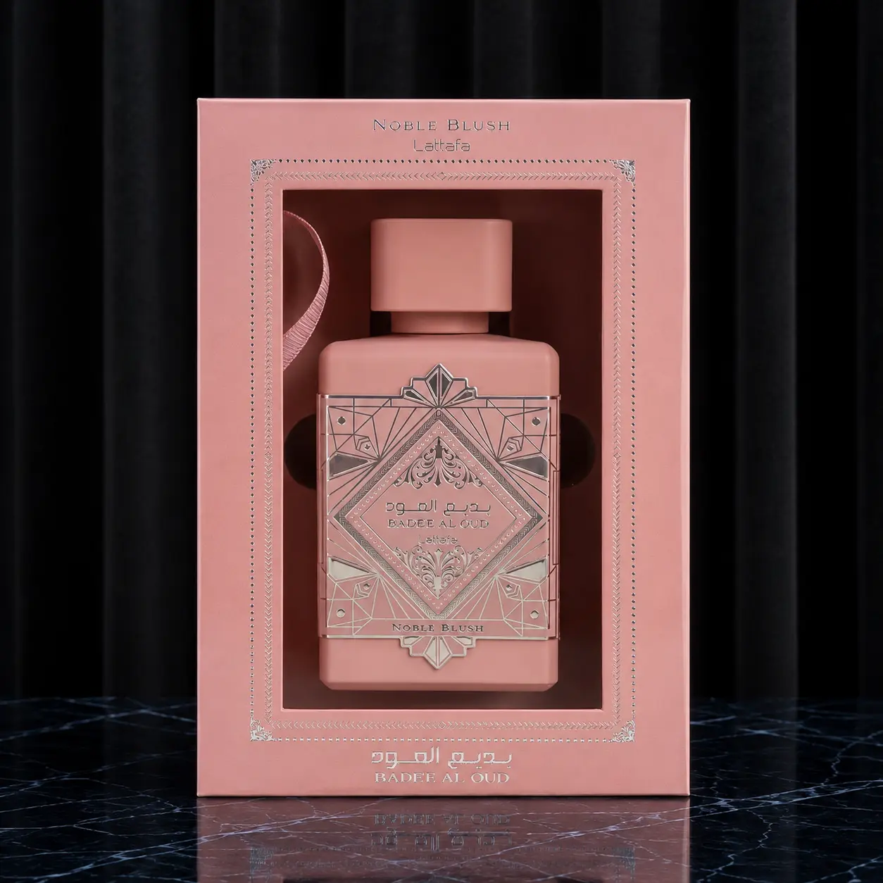 Matte pink display box showcasing the Badee Al Oud Noble Blush bottle inside an ornate silver-bordered frame.