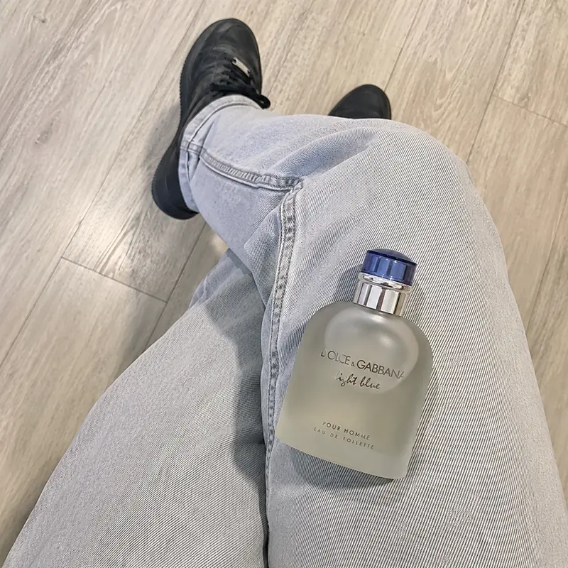 Dolce&Gabbana Light Blue Pour Homme bottle resting on grey denim with black shoes and wood-look flooring.